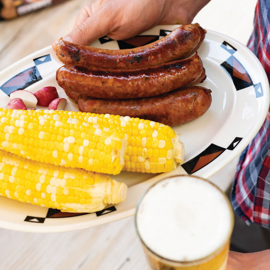 person holding bratwurst on a plate with corn and a beer