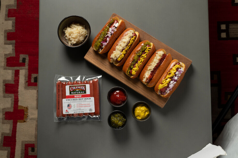 Pack of Coleman hot dogs next two five cooked hot dogs on a wooden plate