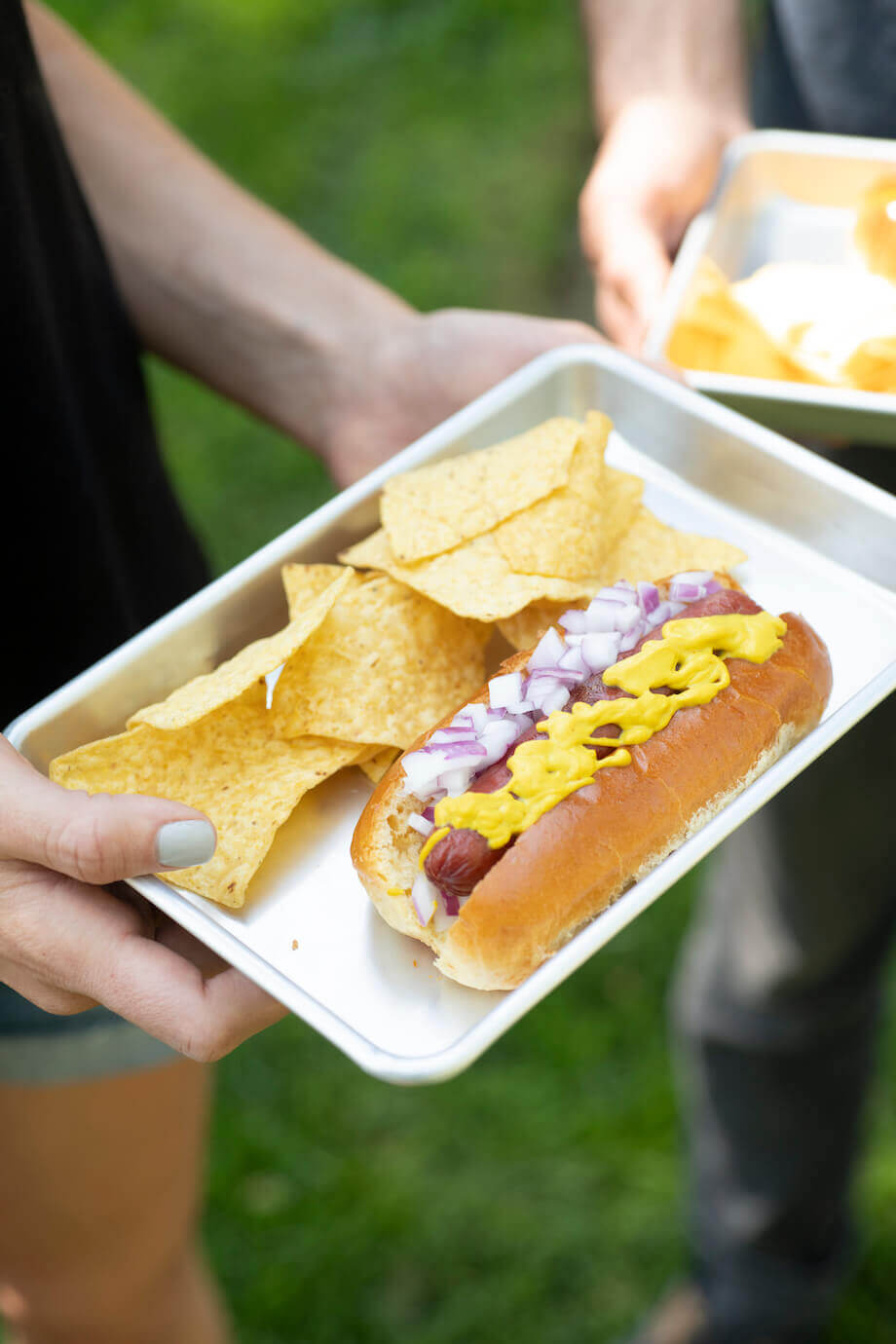 hands holding hot dog tray with chips on the side